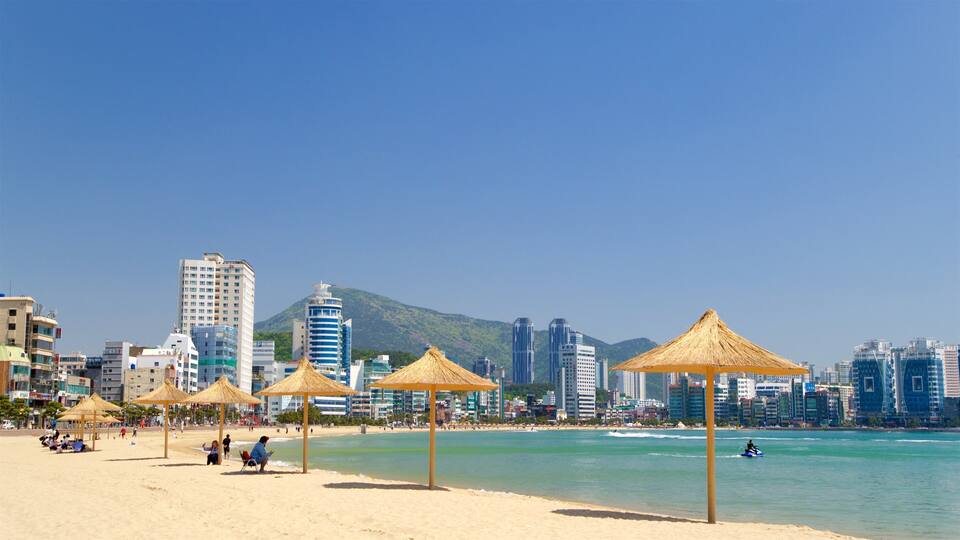 Une plage avec des parasols en chaume, une vue sur les gratte-ciel d’une ville et une montagne en arrière-plan.