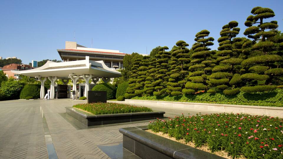 A modern building with a unique white canopy structure, surrounded by well-maintained topiary gardens and flower beds.