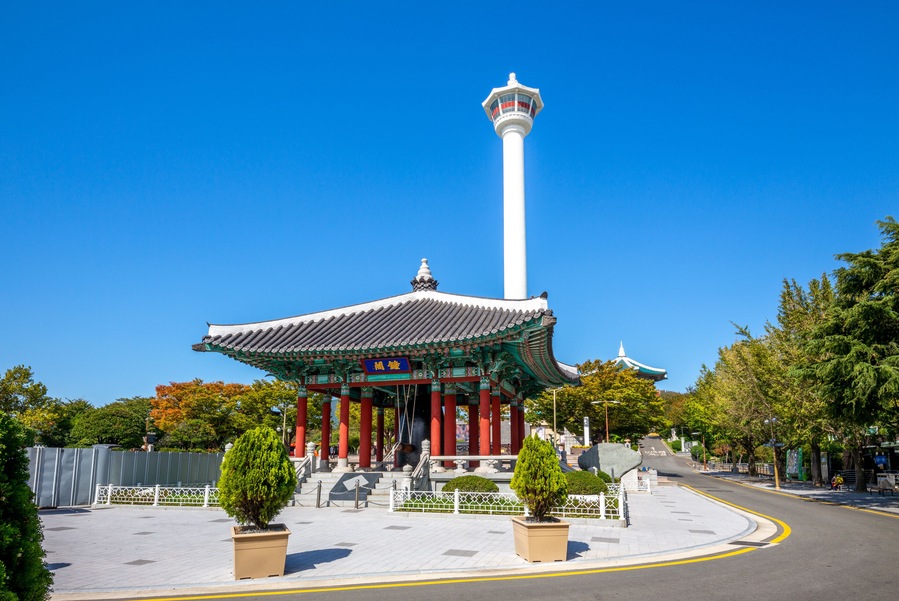 A traditional Korean pavilion with a tall white tower, surrounded by trees and a clear blue sky.
