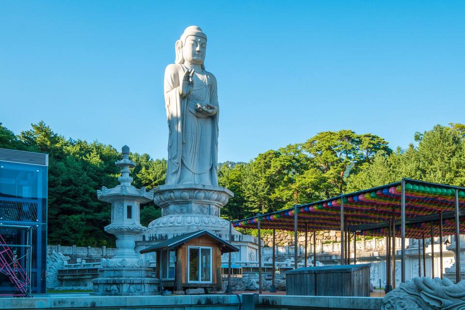 Una gran estatua de piedra de una figura con túnica, una pagoda y un pabellón colorido en un parque.