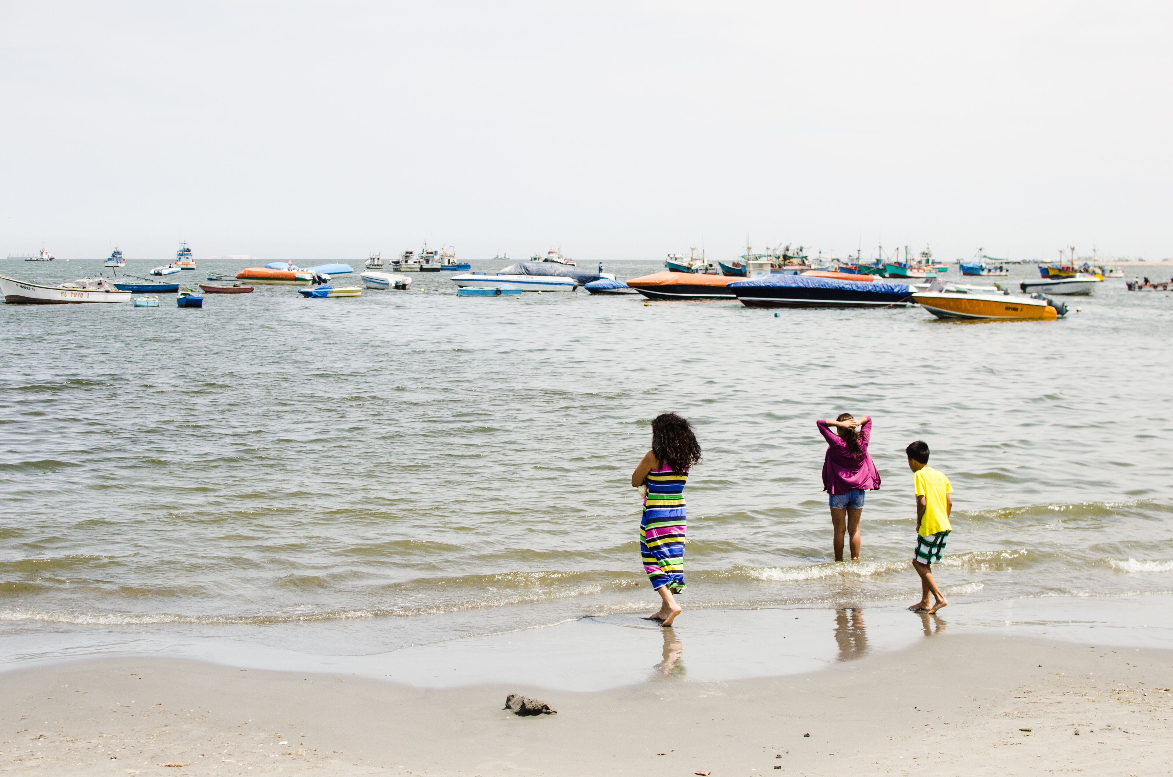 Una playa con gente de pie en aguas poco profundas, y barcos amarrados al fondo.