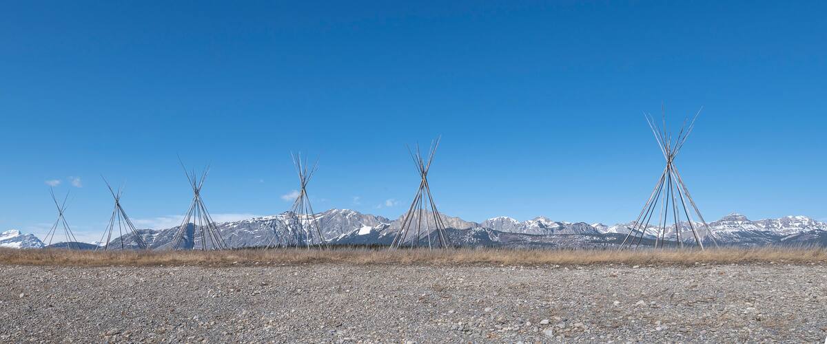 A clear blue sky with a row of traditional Native American tipis in a rocky, grassy plain with snow-capped mountains in the background.