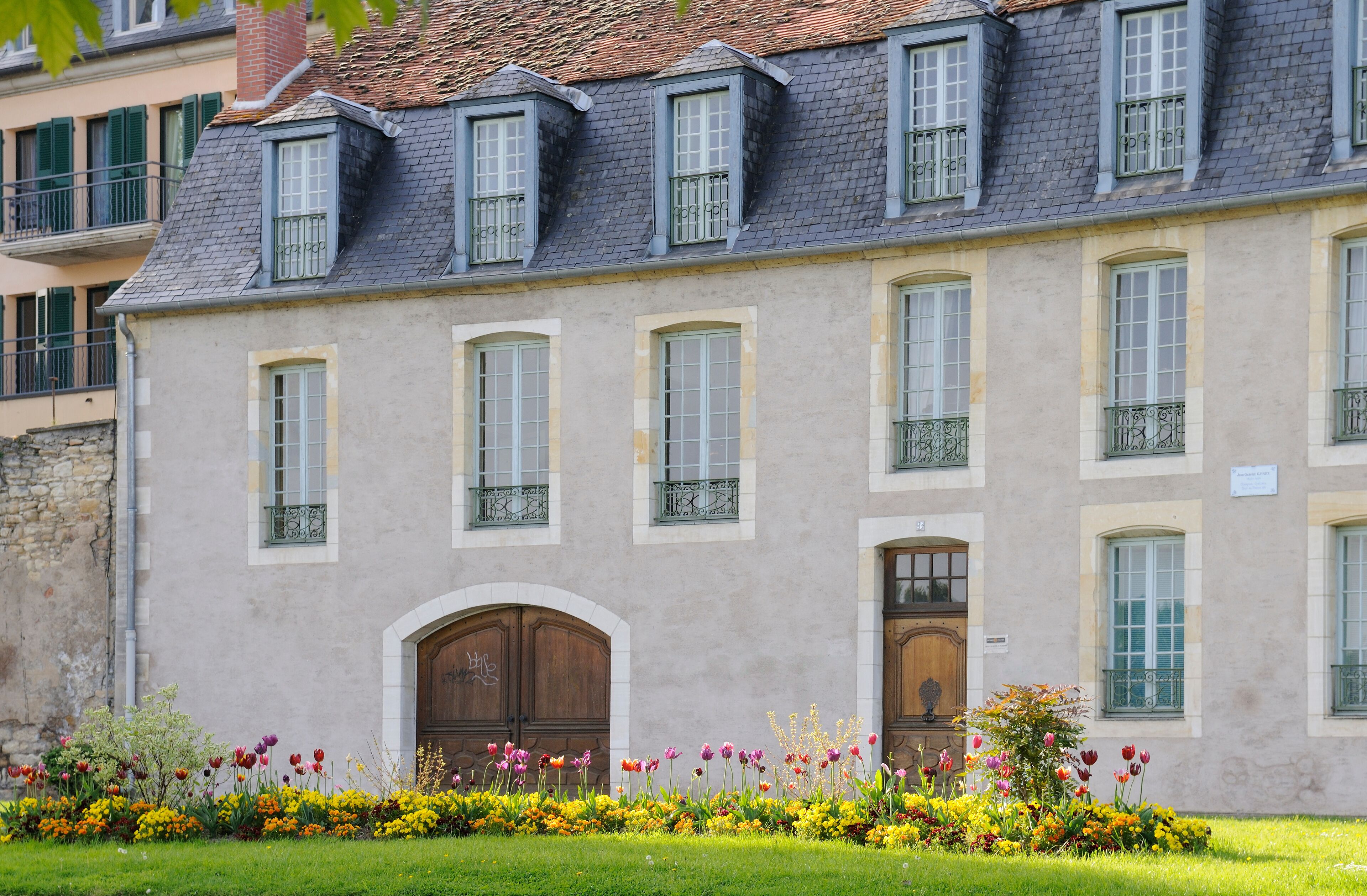 A historic building with a grey facade, multiple windows, and a flower garden in front.