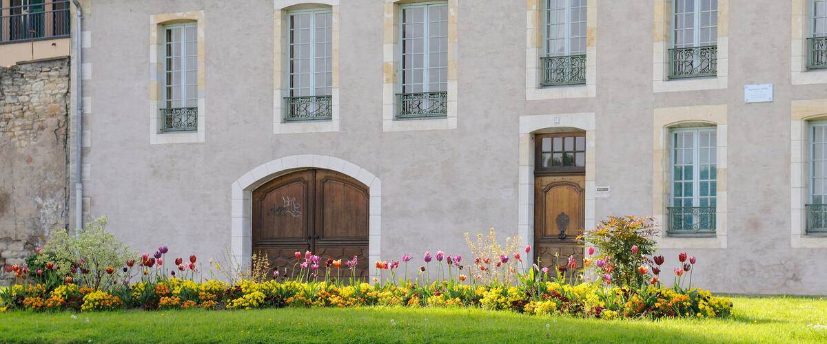A historic building with a grey facade, multiple windows, and a flower garden in front.