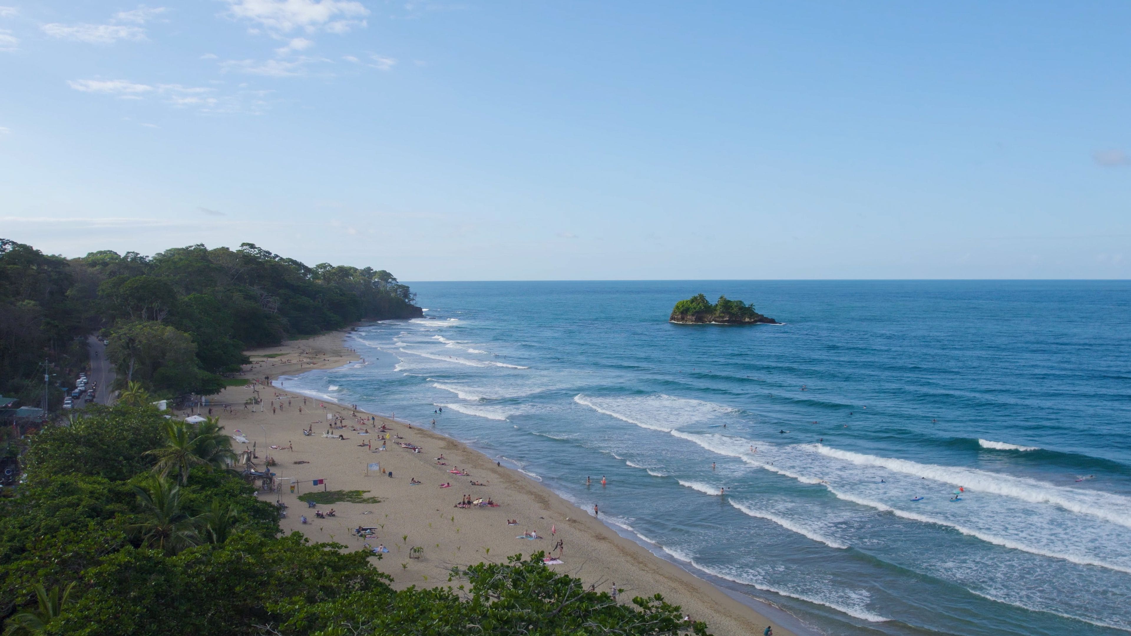 Une plage où des gens se prélassent au soleil et se baignent, une petite île à l’horizon et une côte bordée d’une forêt.