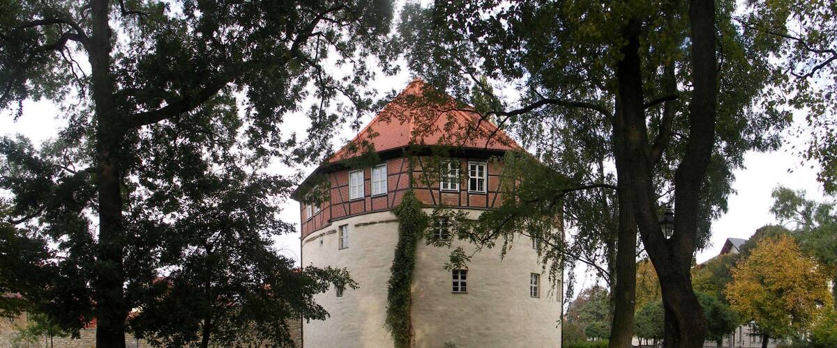 A historic building with a red-tiled roof, surrounded by trees and a grassy area.