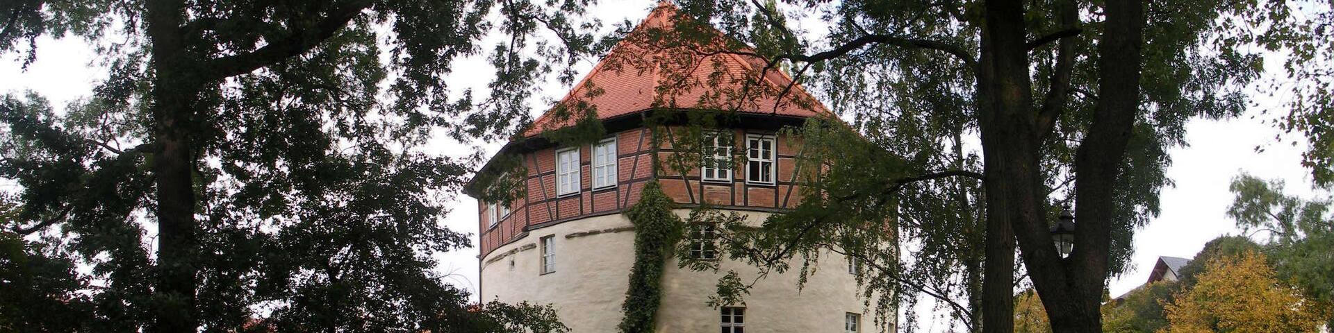 A historic building with a red-tiled roof, surrounded by trees and a grassy area.