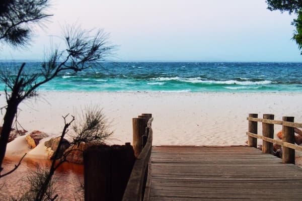 A wooden pier leading to a sandy beach with turquoise water and rocky outcrops.