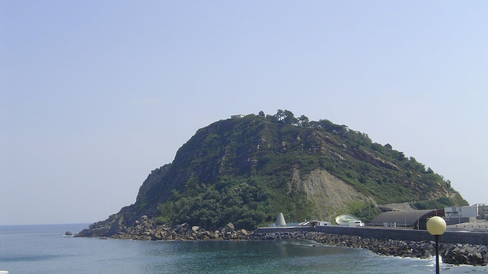 A coastal landscape with a prominent hill covered in greenery, a rocky shoreline, and a clear sky.