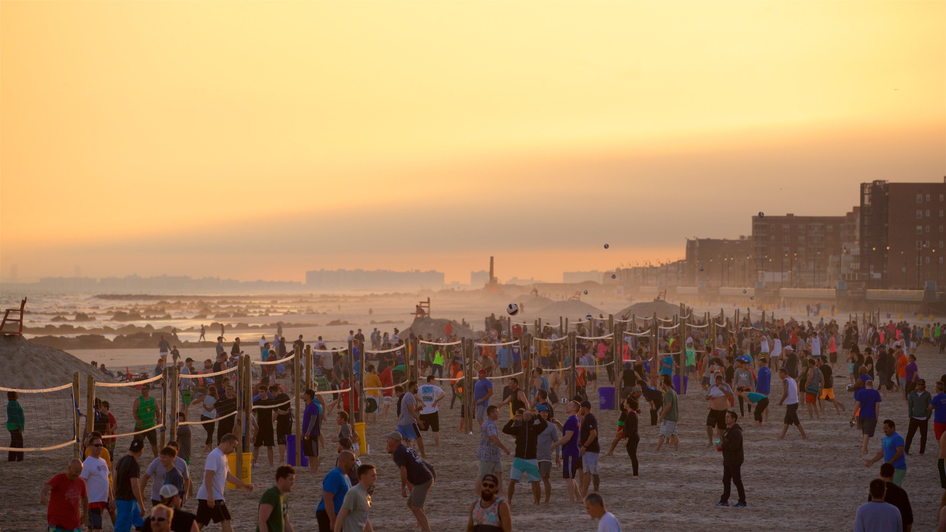 A crowded beach at sunset with people playing volleyball and walking along the shore.