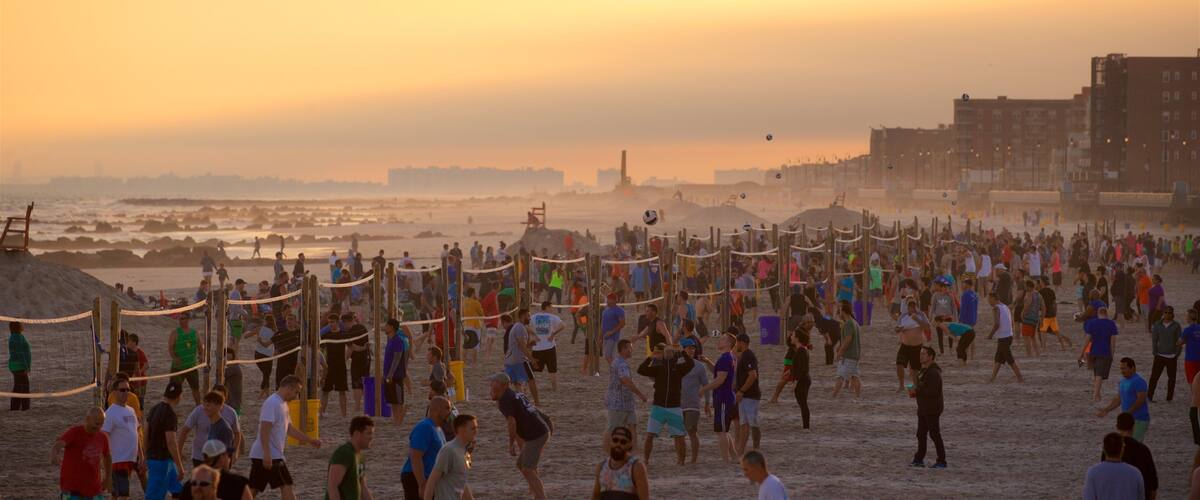A crowded beach at sunset with people playing volleyball and walking along the shore.