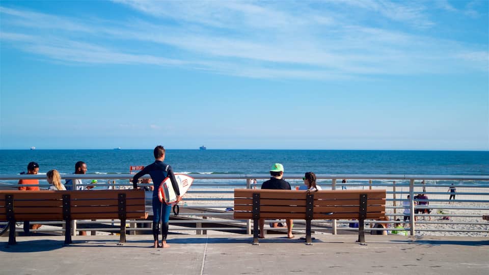 A beach with people sitting on benches, a surfer holding a surfboard, and ships in the distance.