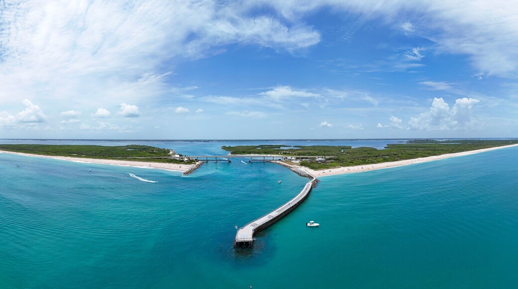 Un paysage côtier avec une longue jetée s’avançant dans des eaux turquoise, une plage de sable et un ciel bleu clair.