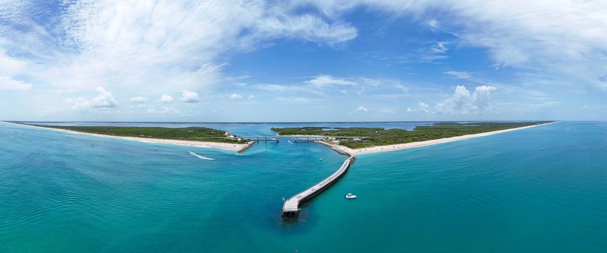 Un paisaje costero con un muelle largo que se adentra en aguas turquesas, una playa arenosa y un cielo azul despejado.