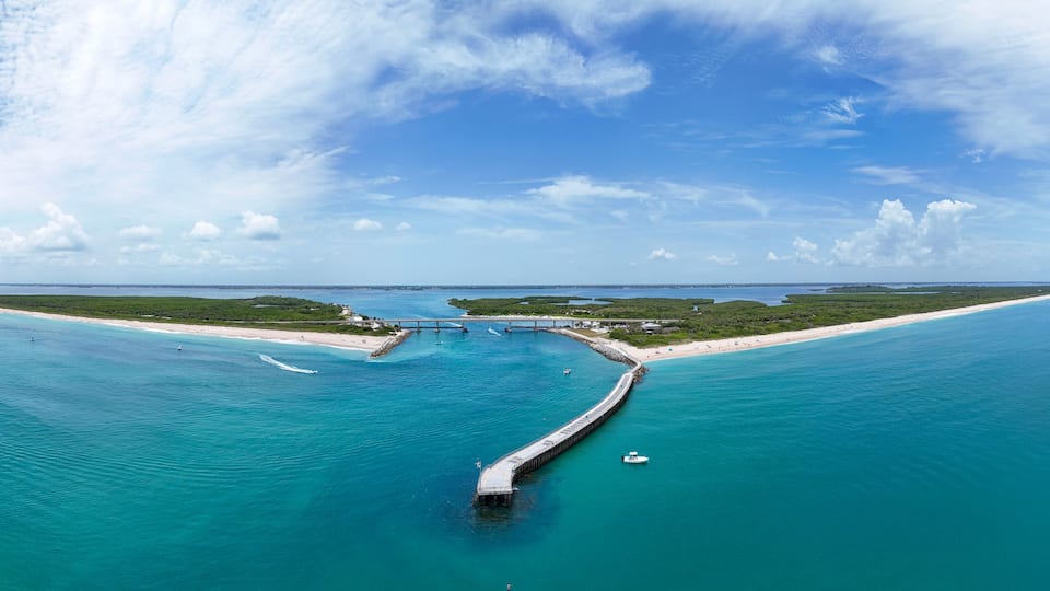 A coastal landscape with a long pier extending into turquoise waters, a sandy beach, and a clear blue sky.