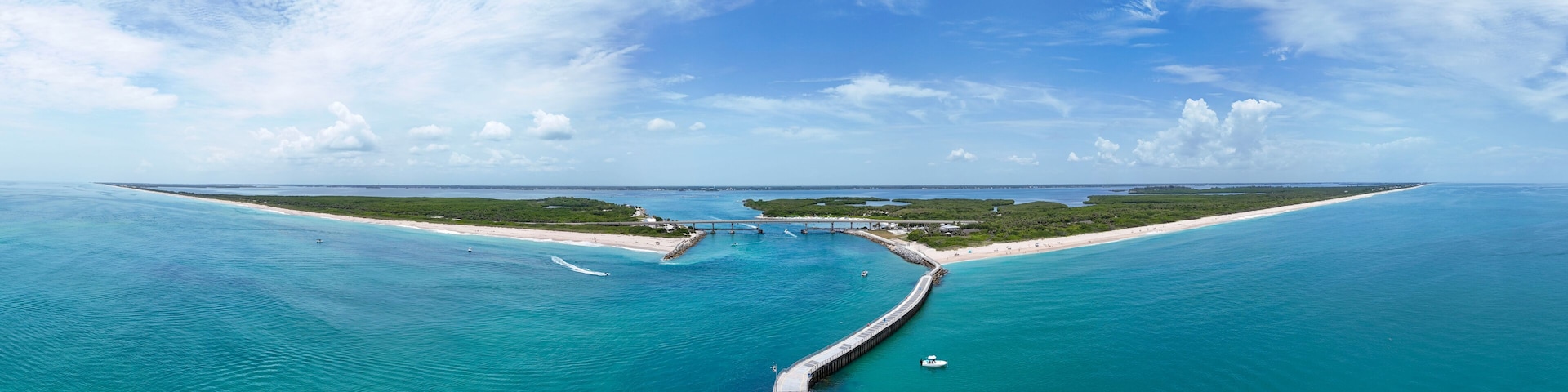 Un paisaje costero con un muelle largo que se adentra en aguas turquesas, una playa arenosa y un cielo azul despejado.