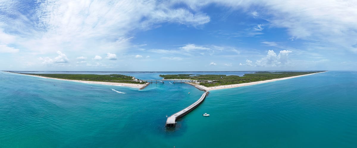 A coastal landscape with a long pier extending into turquoise waters, a sandy beach, and a clear blue sky.