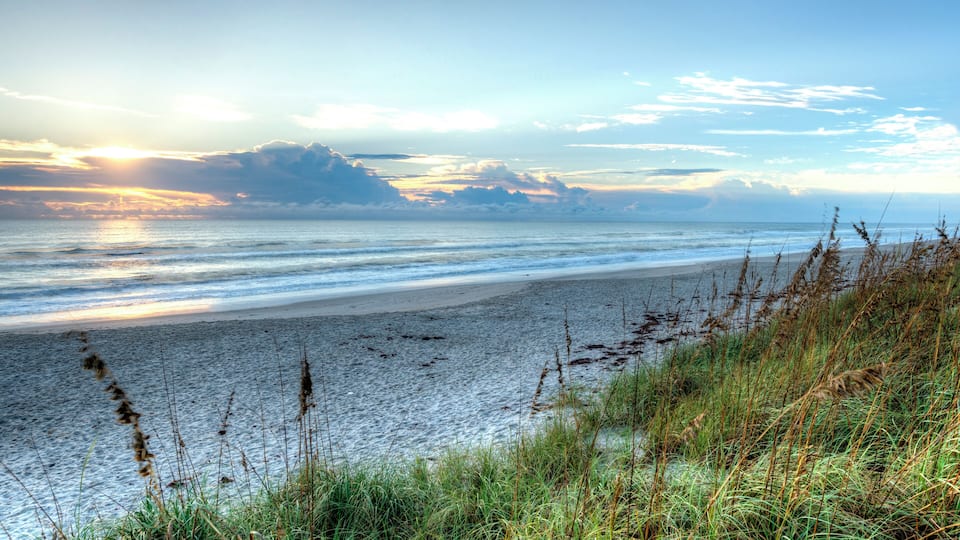 A beach with waves, grassy dunes, and a distant mountain under a clear sky.