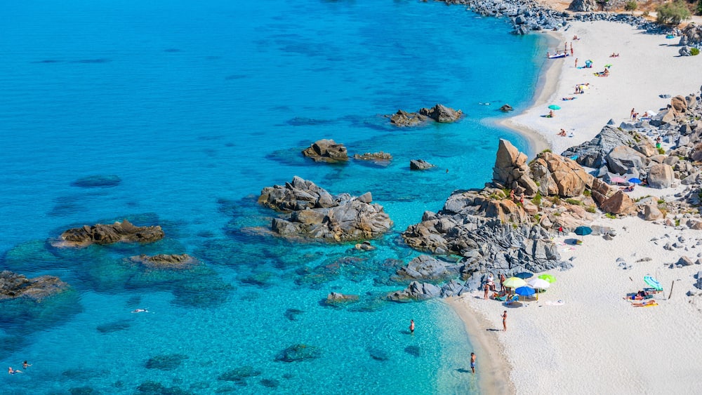 Une plage cĂŽtiĂšre aux eaux turquoise et limpides, parsemĂ©e de rochers et oĂč lâon peut voir des gens se prĂ©lasser au soleil.