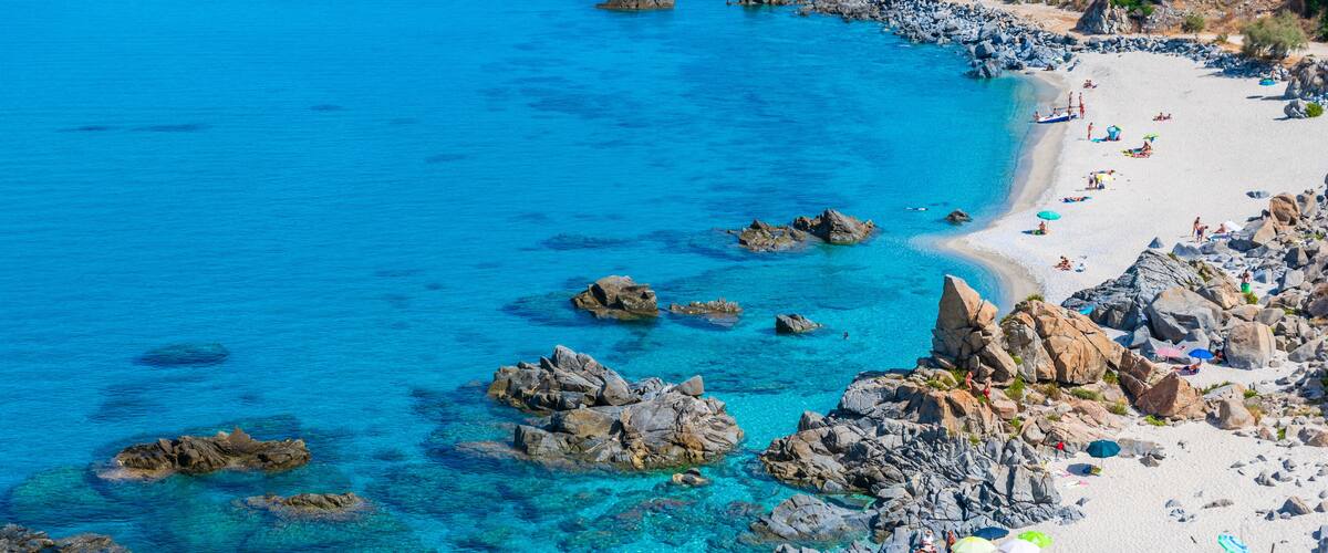 A coastal beach with clear turquoise waters, rocky outcrops, and people sunbathing.