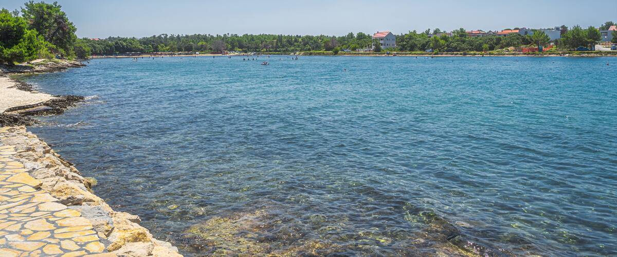 A clear blue lake with a stone walkway along the shore, surrounded by trees and buildings.