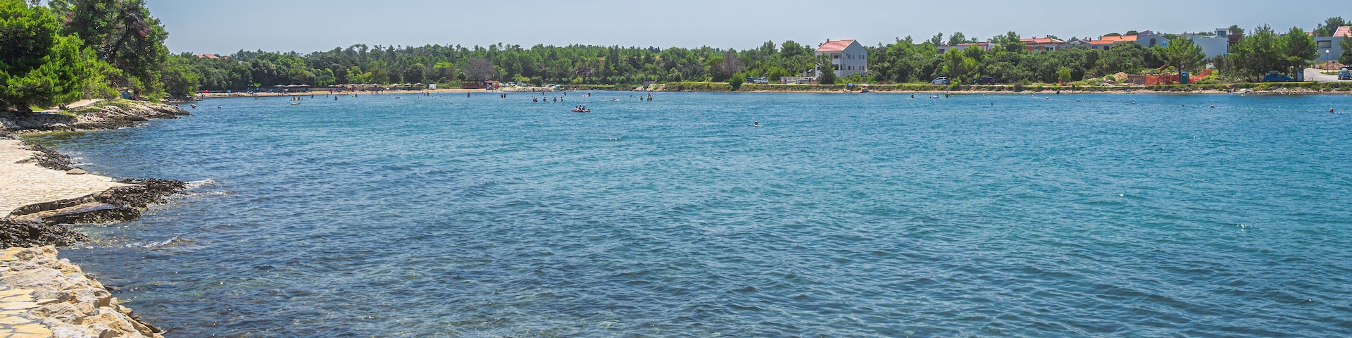 A clear blue lake with a stone walkway along the shore, surrounded by trees and buildings.