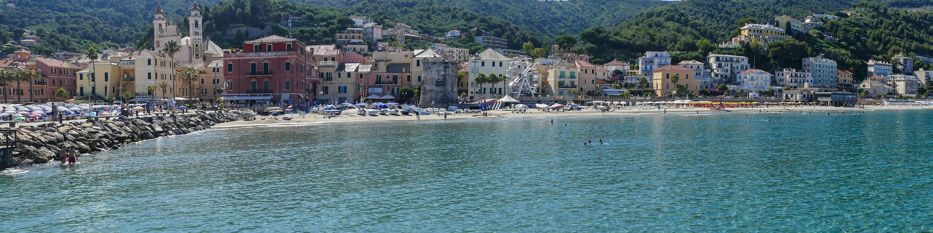 A coastal town with a clear blue sea, a beach with umbrellas, and a hill with a church in the background.