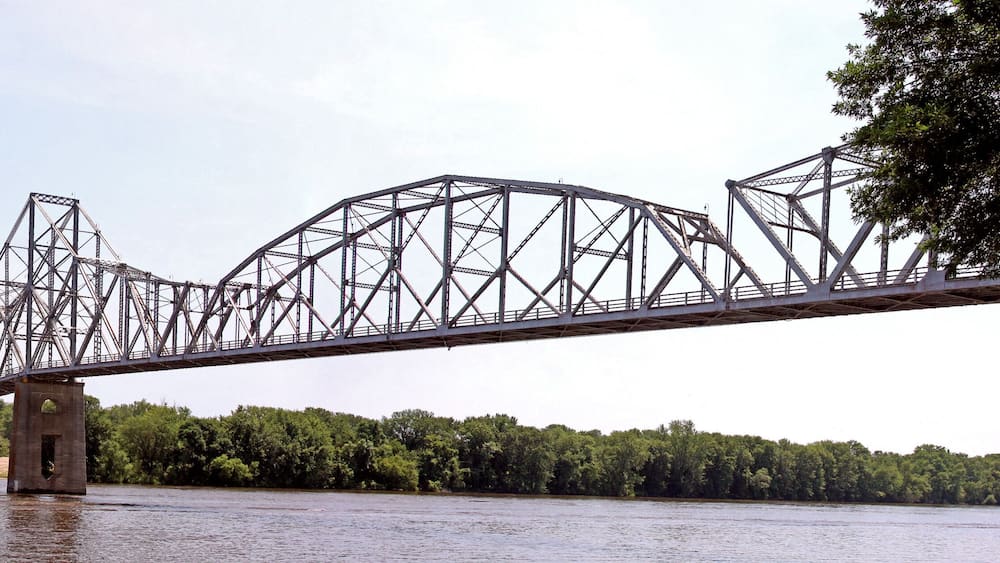 A steel truss bridge over a river with dense green foliage on both sides.