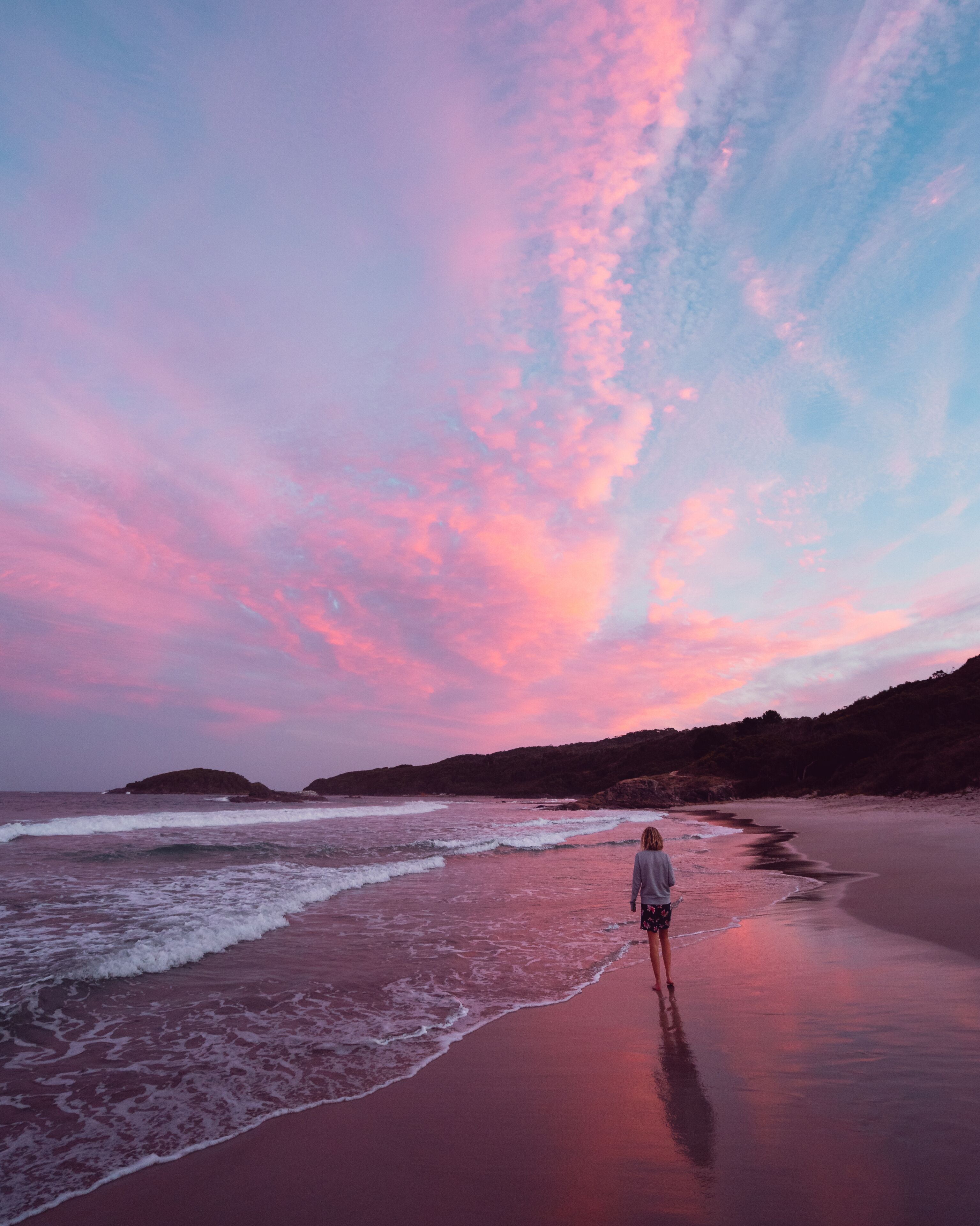 A beach with waves at sunset, a person walking, and distant rocky outcrops.