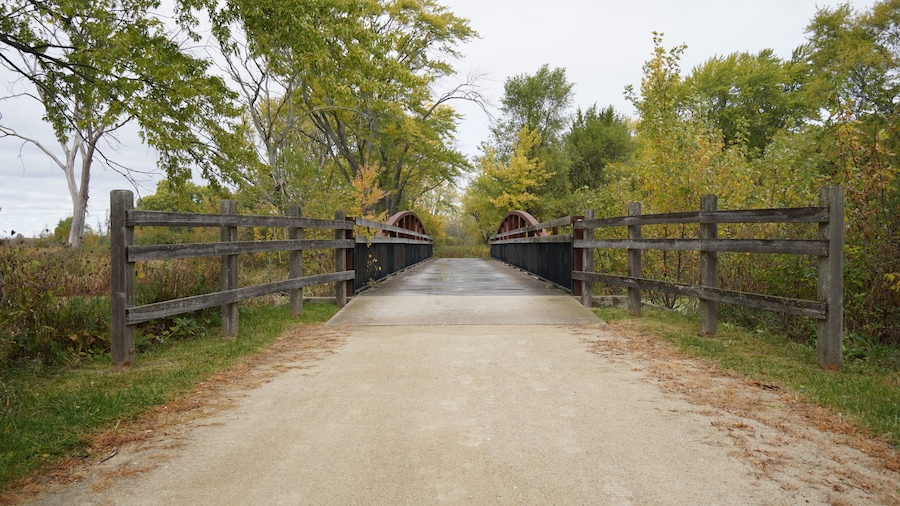 Un puente de madera con arcos metálicos sobre un camino de tierra, rodeado de árboles y follaje.