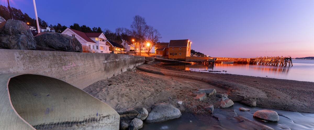 A coastal scene with a concrete structure, a calm beach, and buildings with lights in the background.