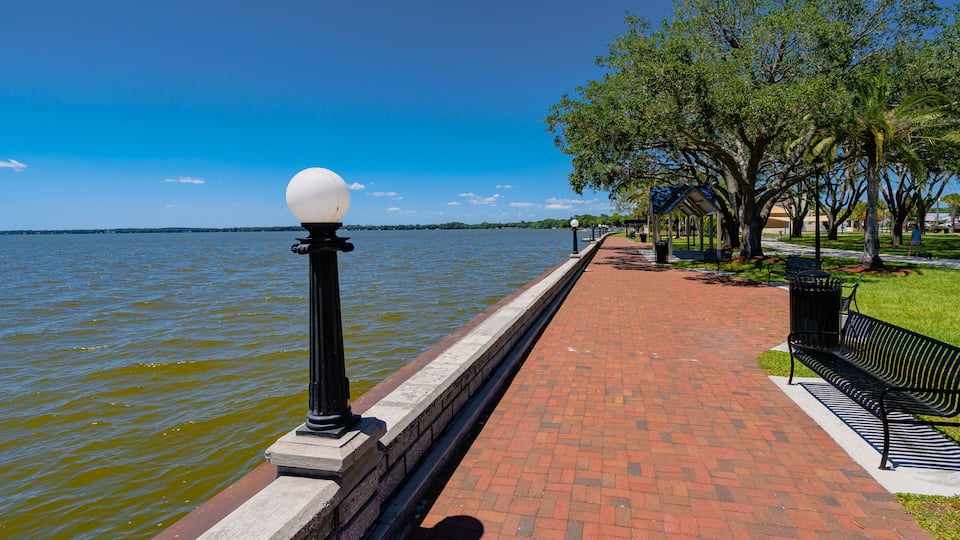 A waterfront walkway with a brick path, black lamp posts, a bench, and trees.