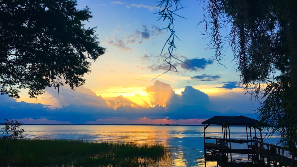 A sunset over a calm lake with a wooden dock and surrounding trees.