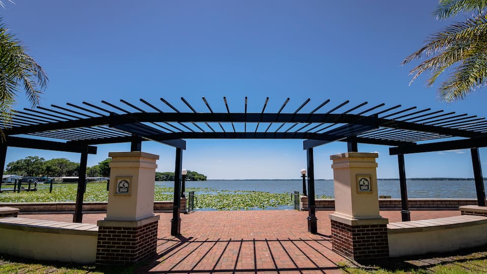 A covered pavilion with a brick floor, palm trees, and a body of water in the background.