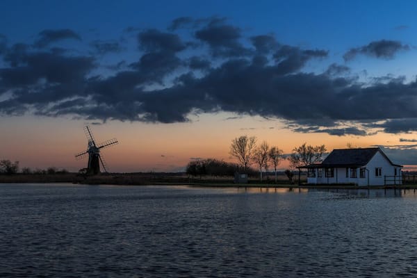 A windmill, a house, and a body of water during sunset.