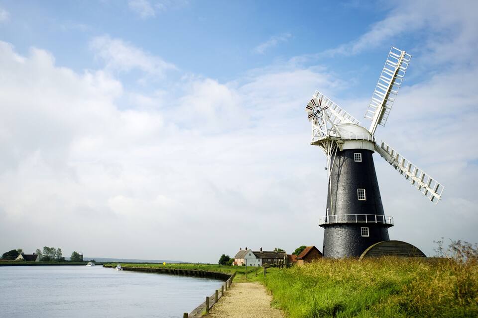 A traditional windmill by a calm river, surrounded by grassy fields and a few houses.