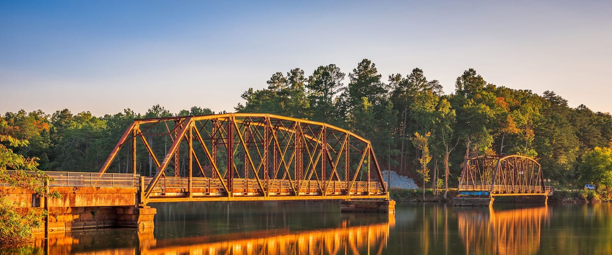 A metal bridge over a calm river with reflections, surrounded by trees and a clear sky.