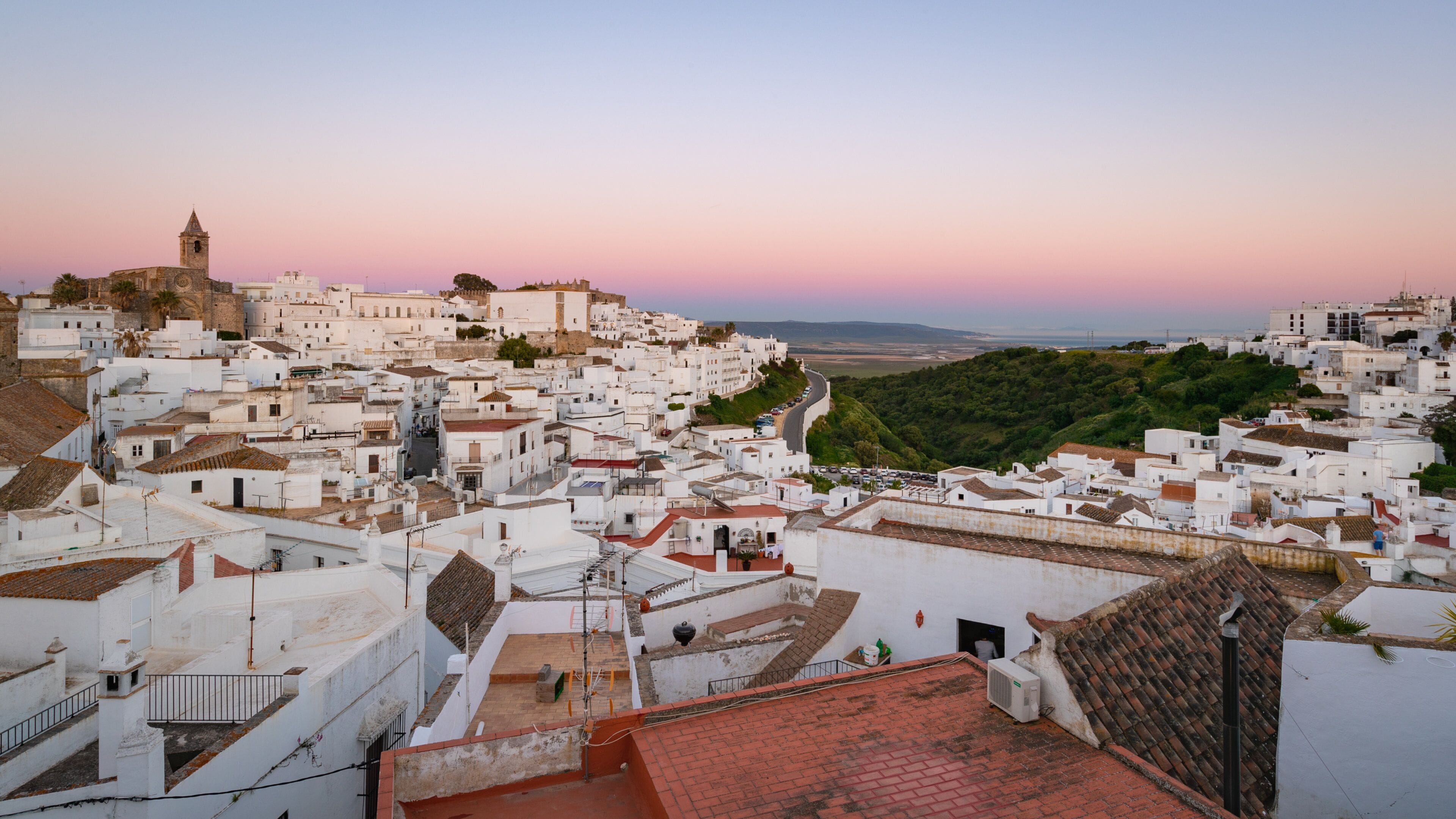 A coastal town with white buildings, a prominent church tower, and a hilly backdrop.