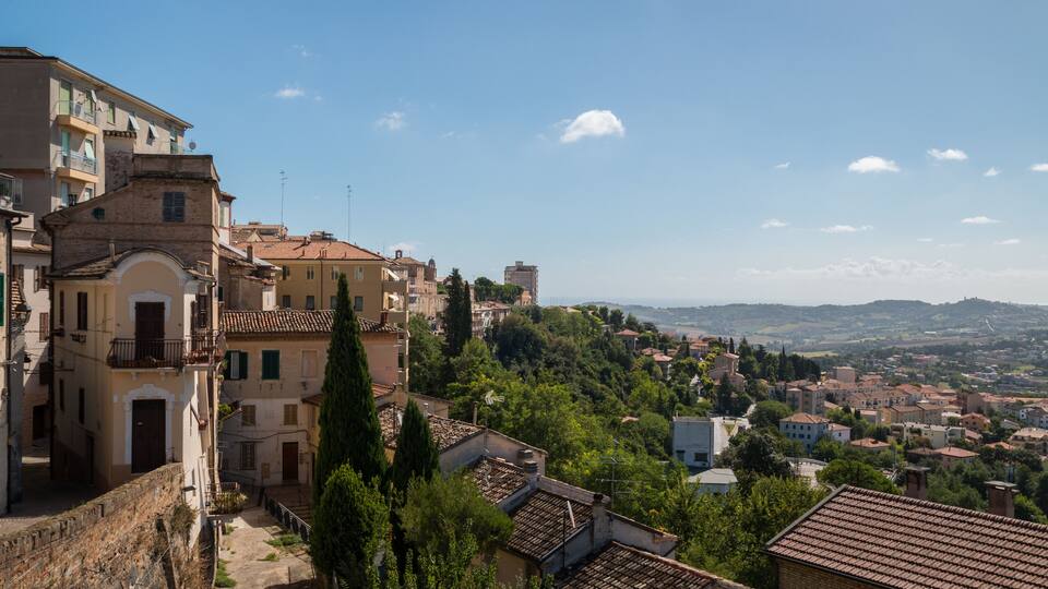 A hillside town with terracotta-roofed buildings, a stone wall, and a view of distant hills.