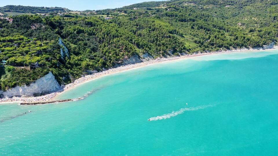 A turquoise coastal beach with a rocky shoreline, dense forested hills in the background, and a clear sky.