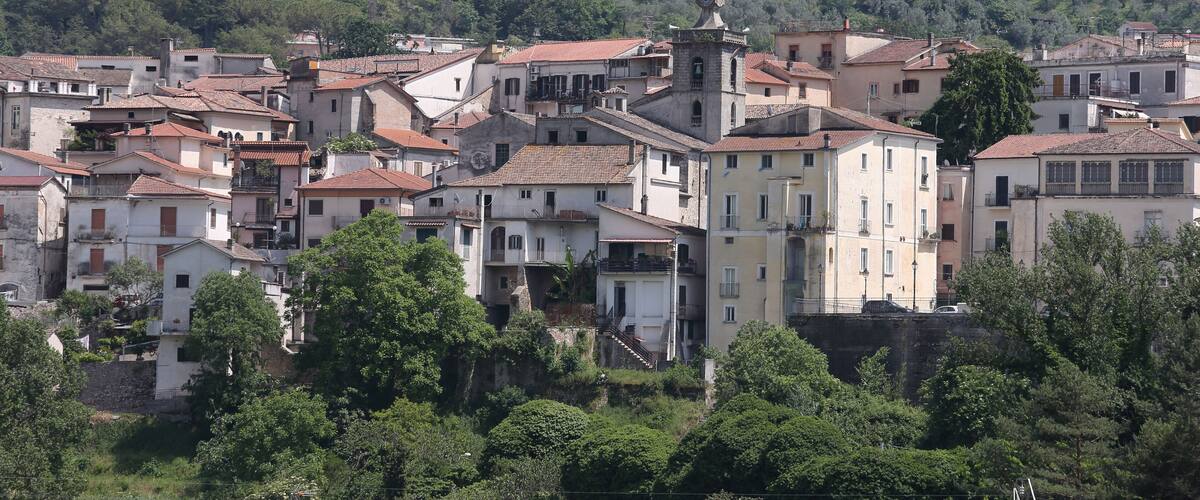 A hillside town with terracotta-roofed buildings, a prominent tower, and lush green trees.
