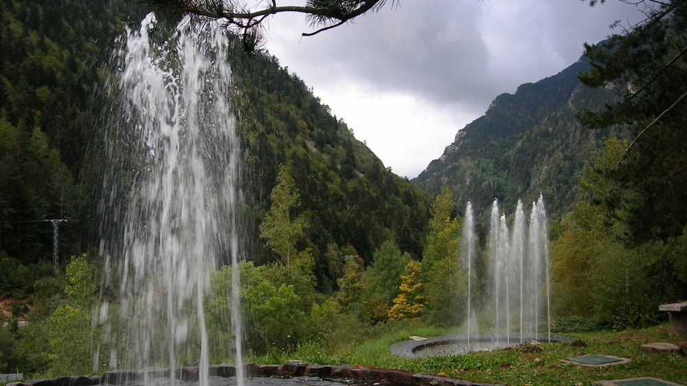 A mountainous landscape with multiple waterfalls cascading into a pond surrounded by lush greenery and pine trees.