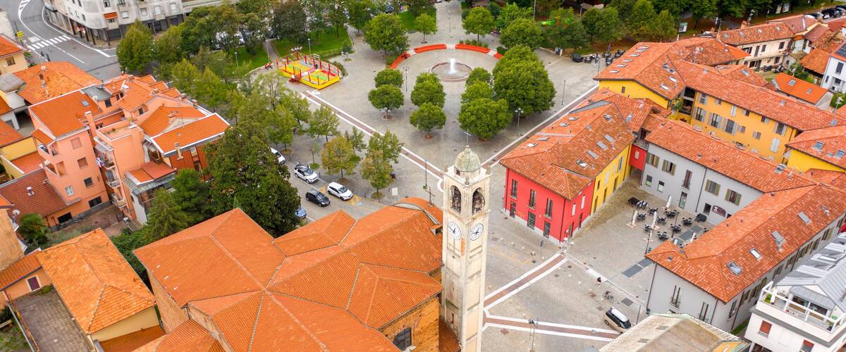 An aerial view of a town square with a prominent clock tower, surrounded by colorful buildings with red-tiled roofs.