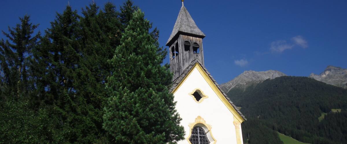 A small white church with a bell tower, surrounded by lush green trees and mountains in the background.