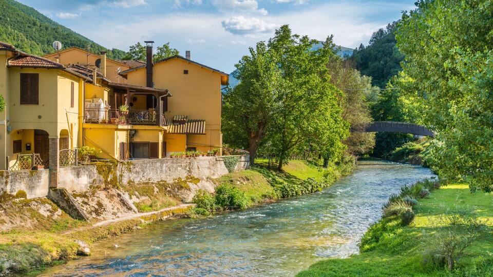 A river flowing through a lush valley with a bridge, surrounded by greenery and a yellow building with a balcony.