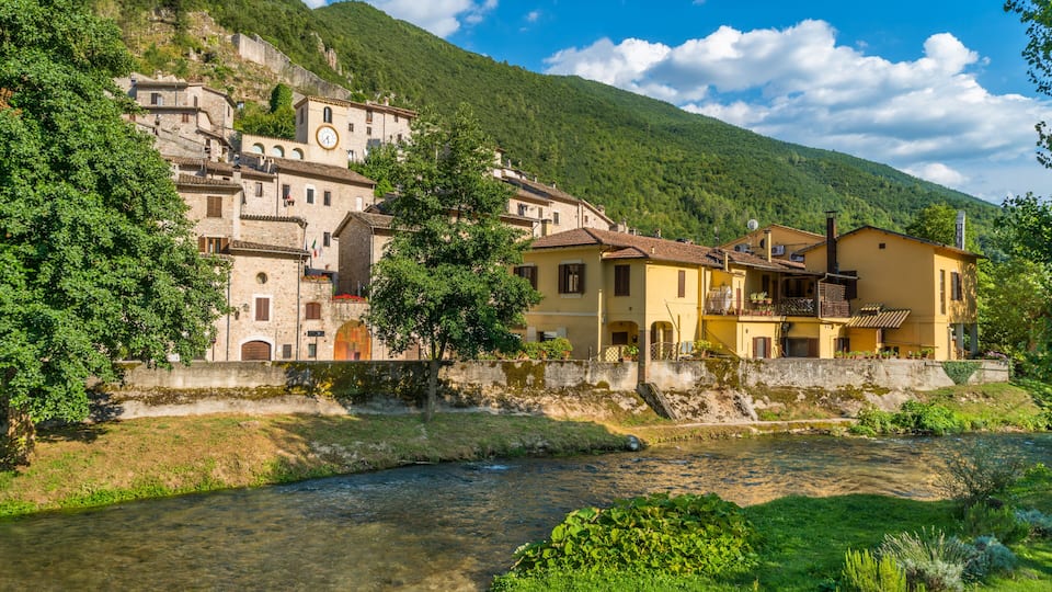 A river flowing through a village with historic buildings and a mountain in the background.