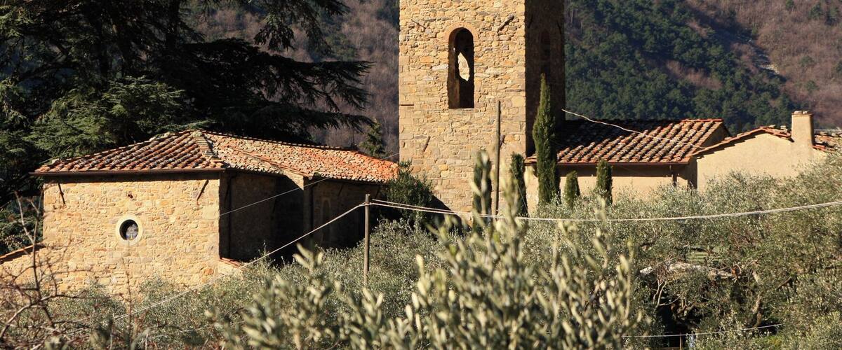 A stone church with a bell tower and tiled roofs, surrounded by mountains and cypress trees.