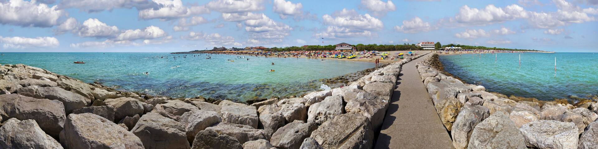 A rocky shoreline with clear turquoise water, a paved walkway, and a beach with umbrellas and people in the distance.
