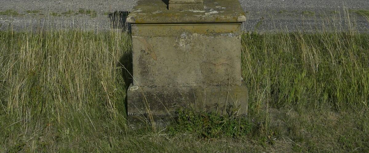 A stone cross with a crucifixion scene in the center, set against a rural landscape with fields and a clear sky.