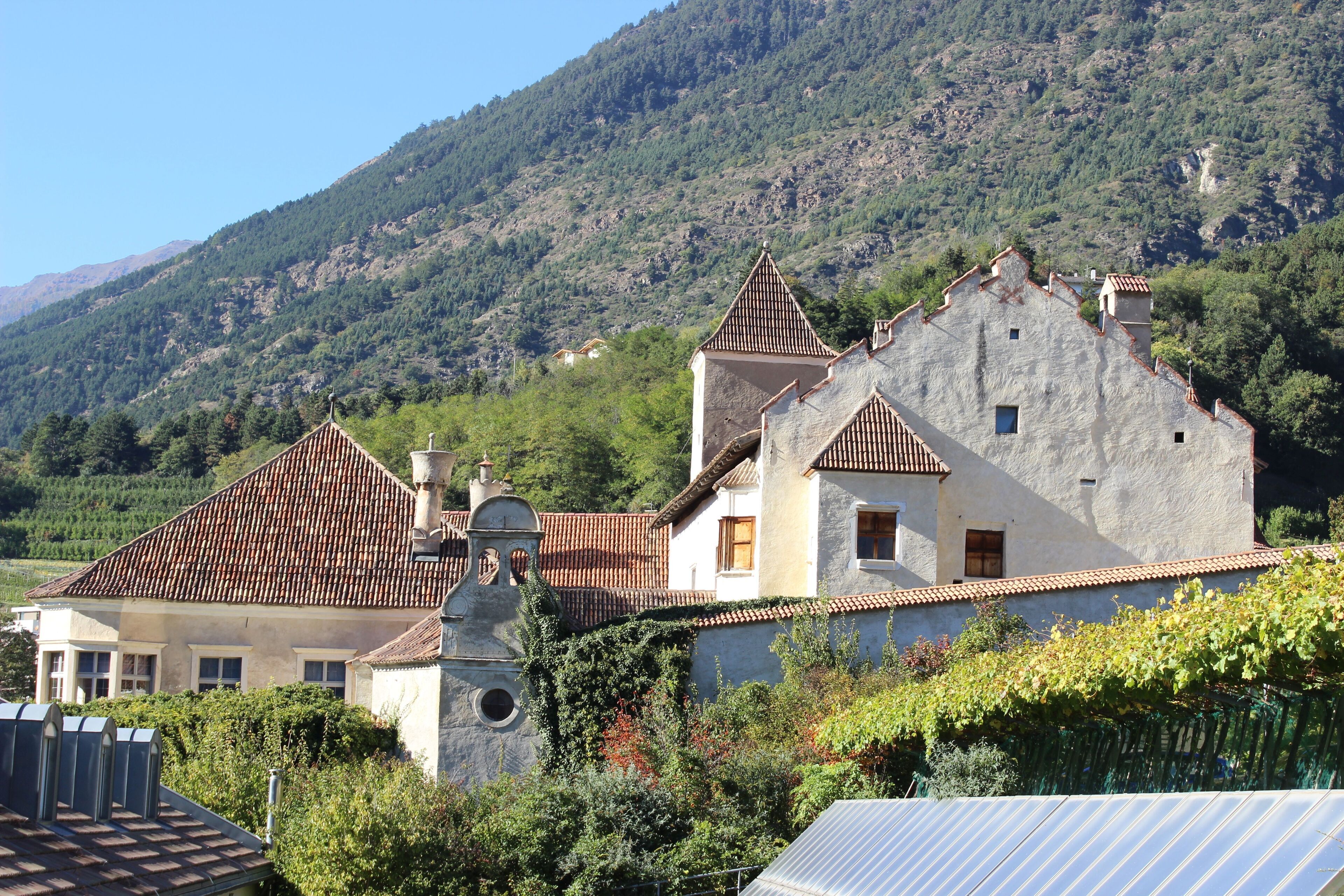 A traditional stone house with a tiled roof, surrounded by lush greenery and a mountainous backdrop.